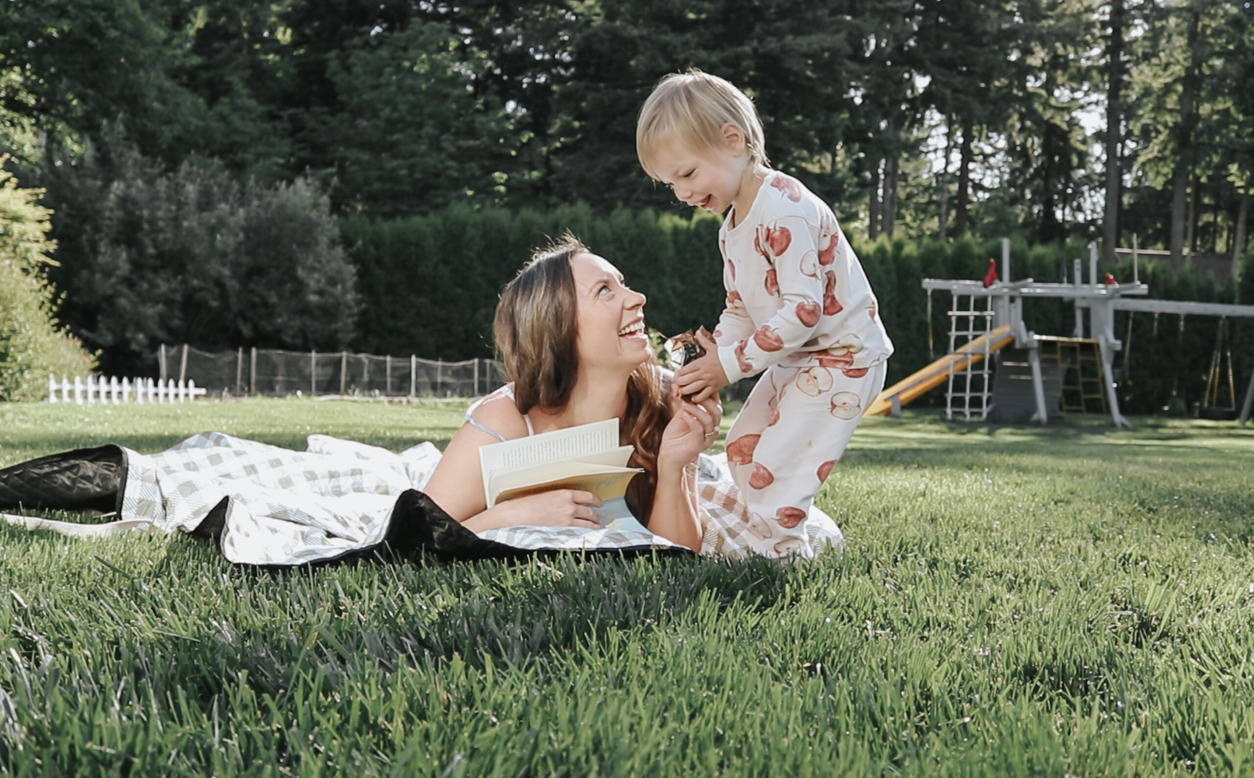 Christian mom lays on a blanked on the lawn with a book in her hand playing with her laughing child